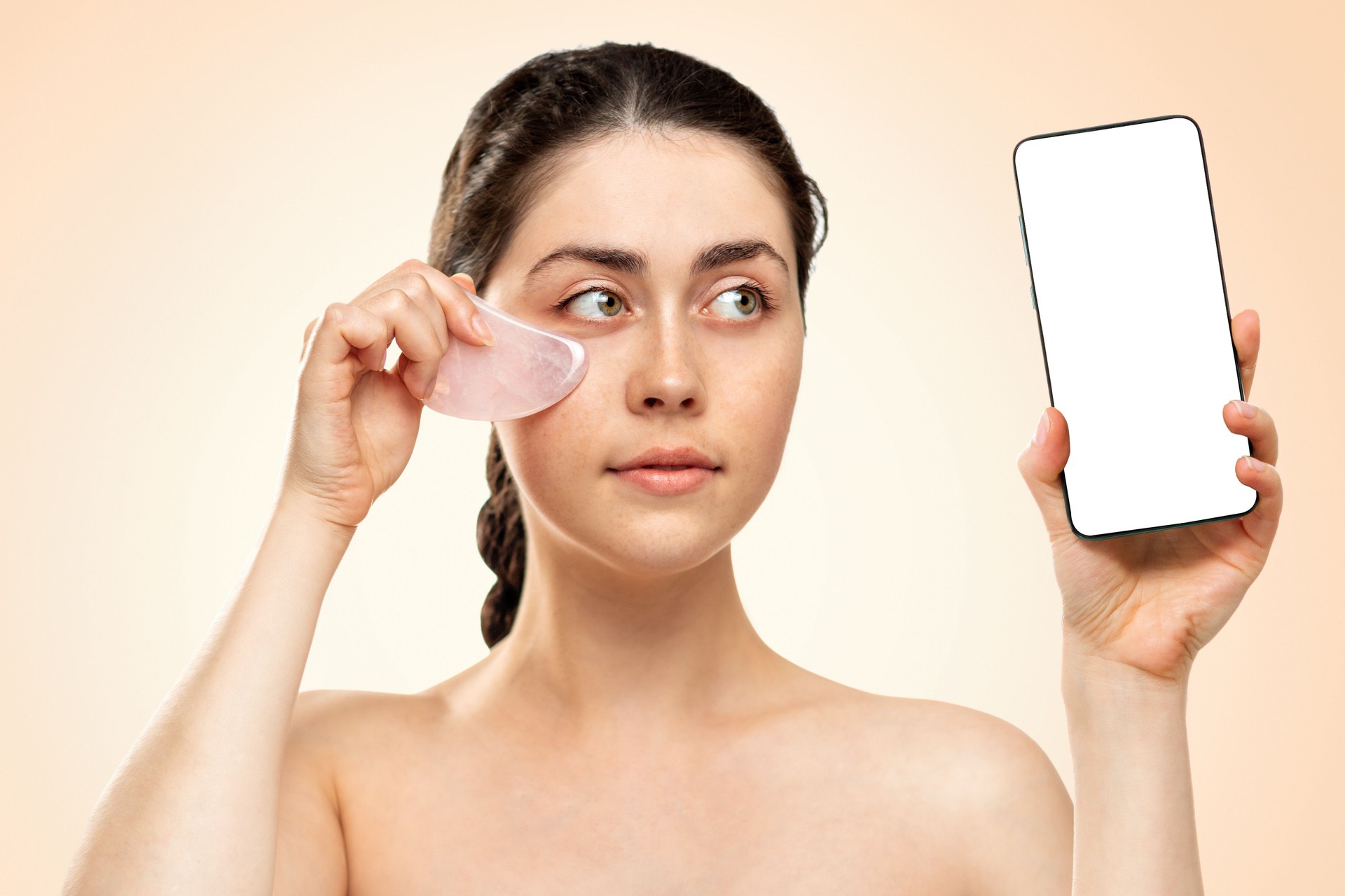 Massage Gua Sha. Une jeune femme tient un téléphone portable dans une main et une pierre de quartz gua Sha dans l’autre. Maquette. Fond beige. Concept d’entreprise et de cosmétologie
